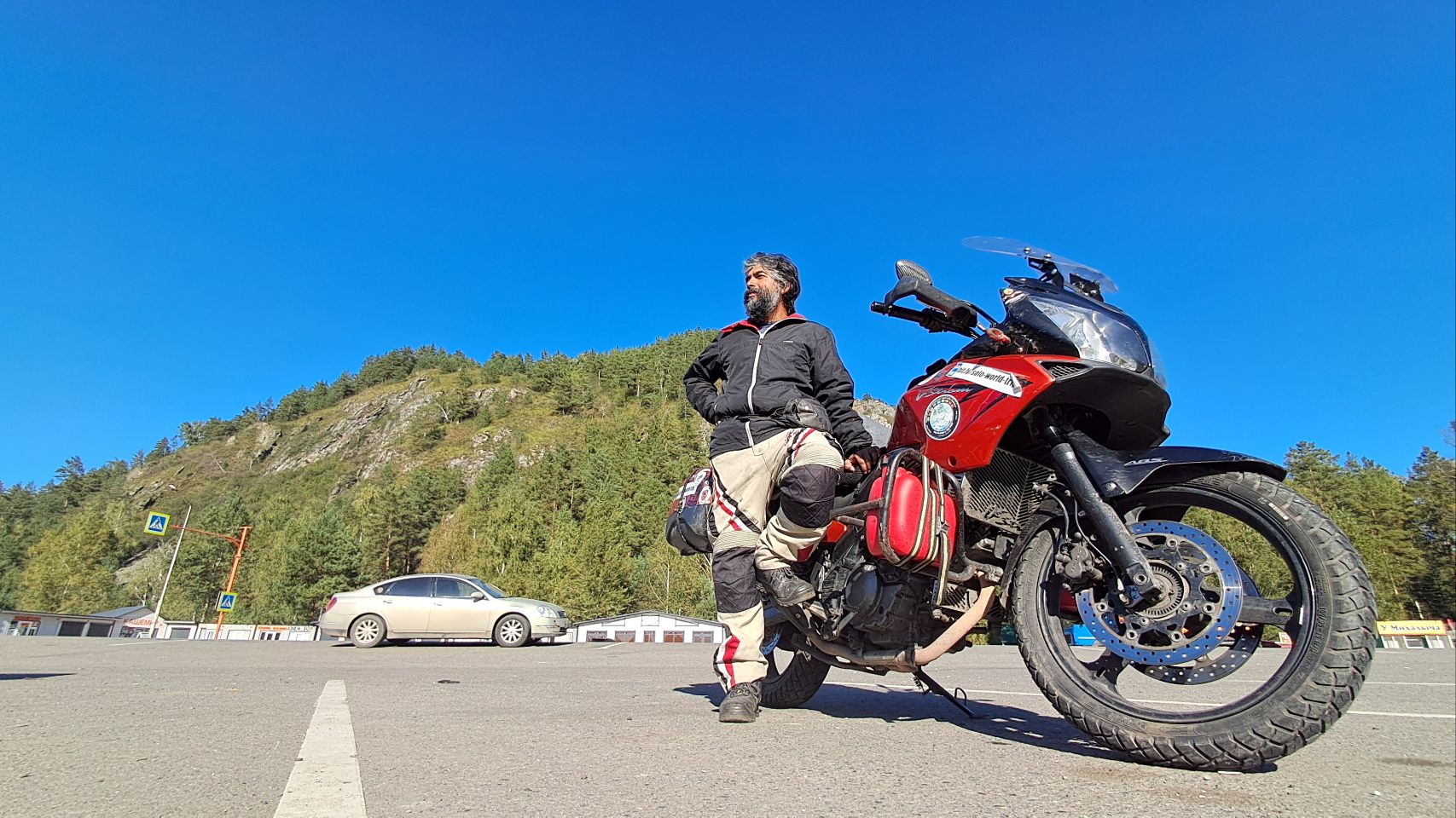 Ashish with his red adventure motorcycle on a road in front of a forested hill under a clear blue sky